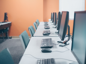 A row of desktop computers on desks with chairs.