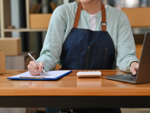 Small business owner in apron writing notes on a clipboard while working on a laptop at a desk