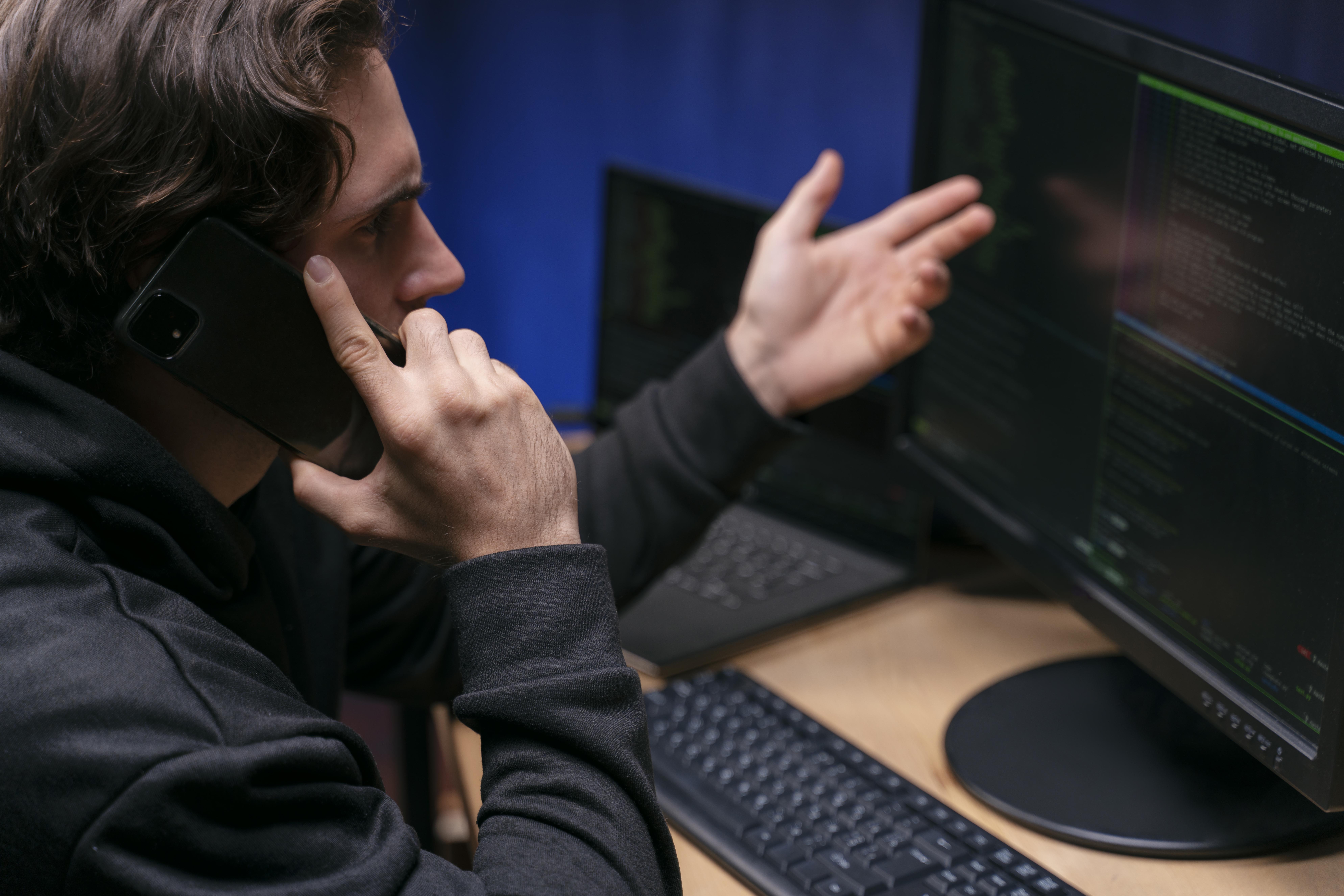 A man in a dark hoodie talks on the phone while gesturing toward a computer screen displaying lines of code