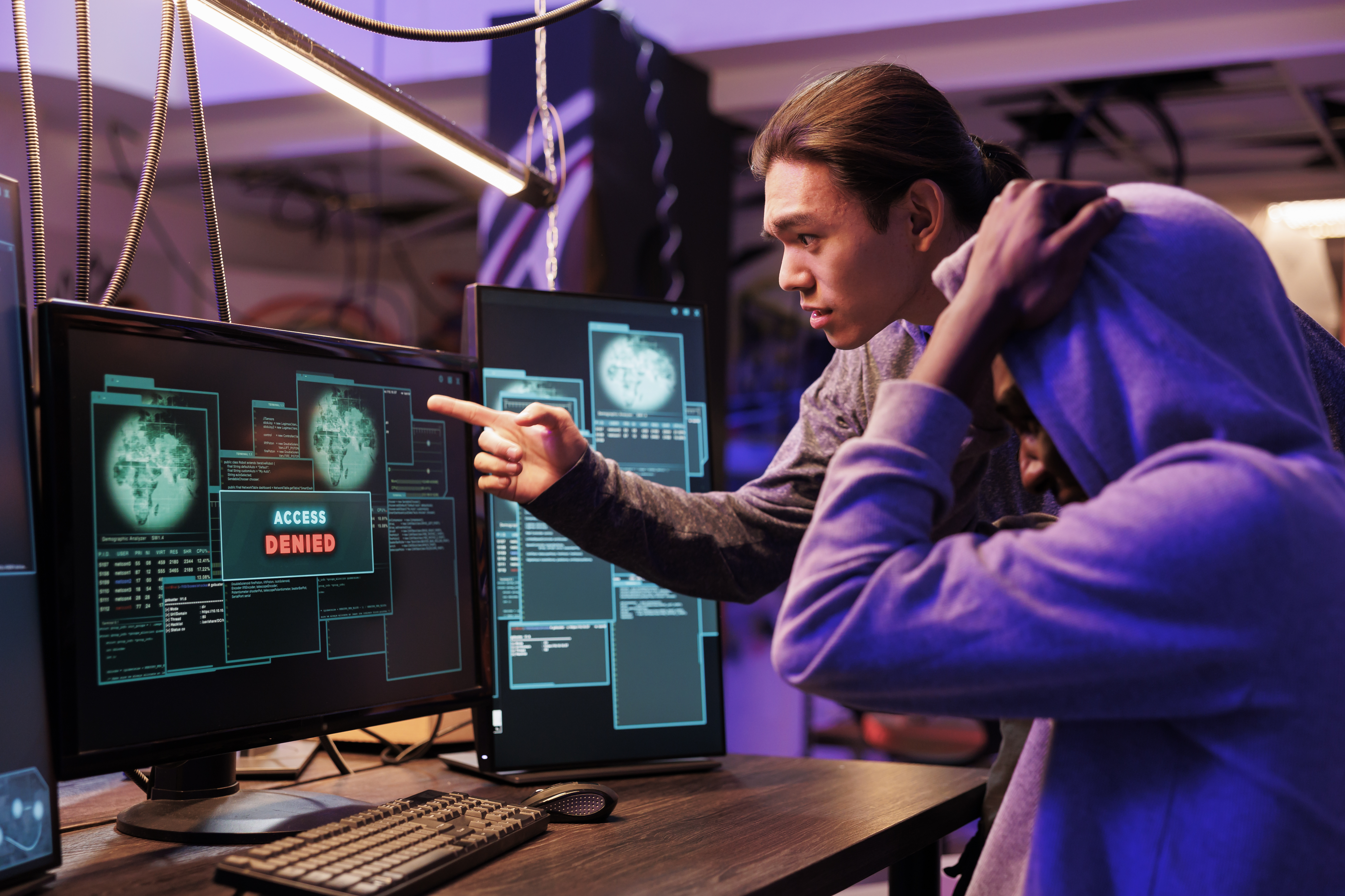 Two people in a dimly lit room looking at computer screens displaying an “Access Denied” message, one pointing at the monitor while the other holds their head in frustration.