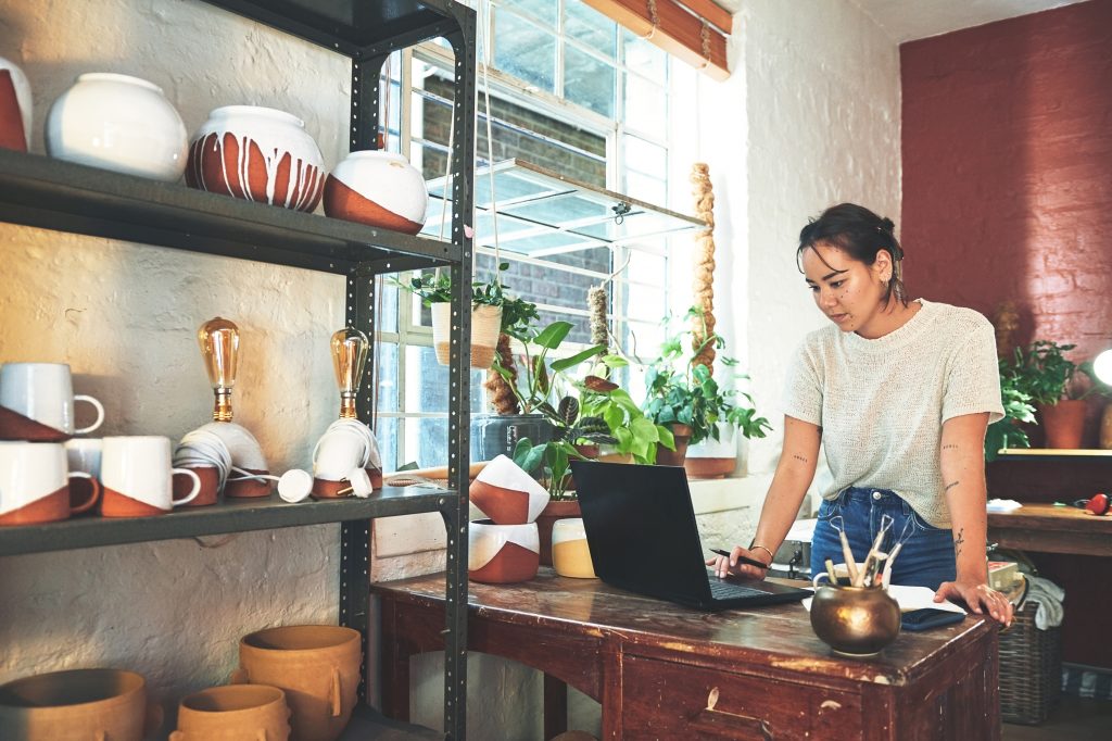 Small business owner in a pottery studio reviewing inventory and orders on a laptop surrounded by handmade ceramics.