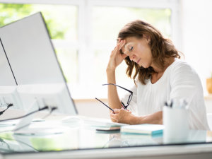 Stressed businesswoman sitting at a desk with her hand on her forehead, holding glasses, looking frustrated in front of a computer monitor in a bright office setting.