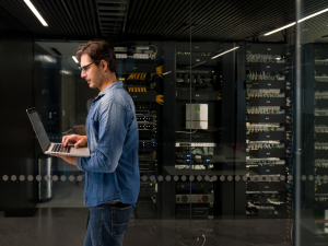 IT technician working on a laptop inside a server room monitoring small business technology systems.
