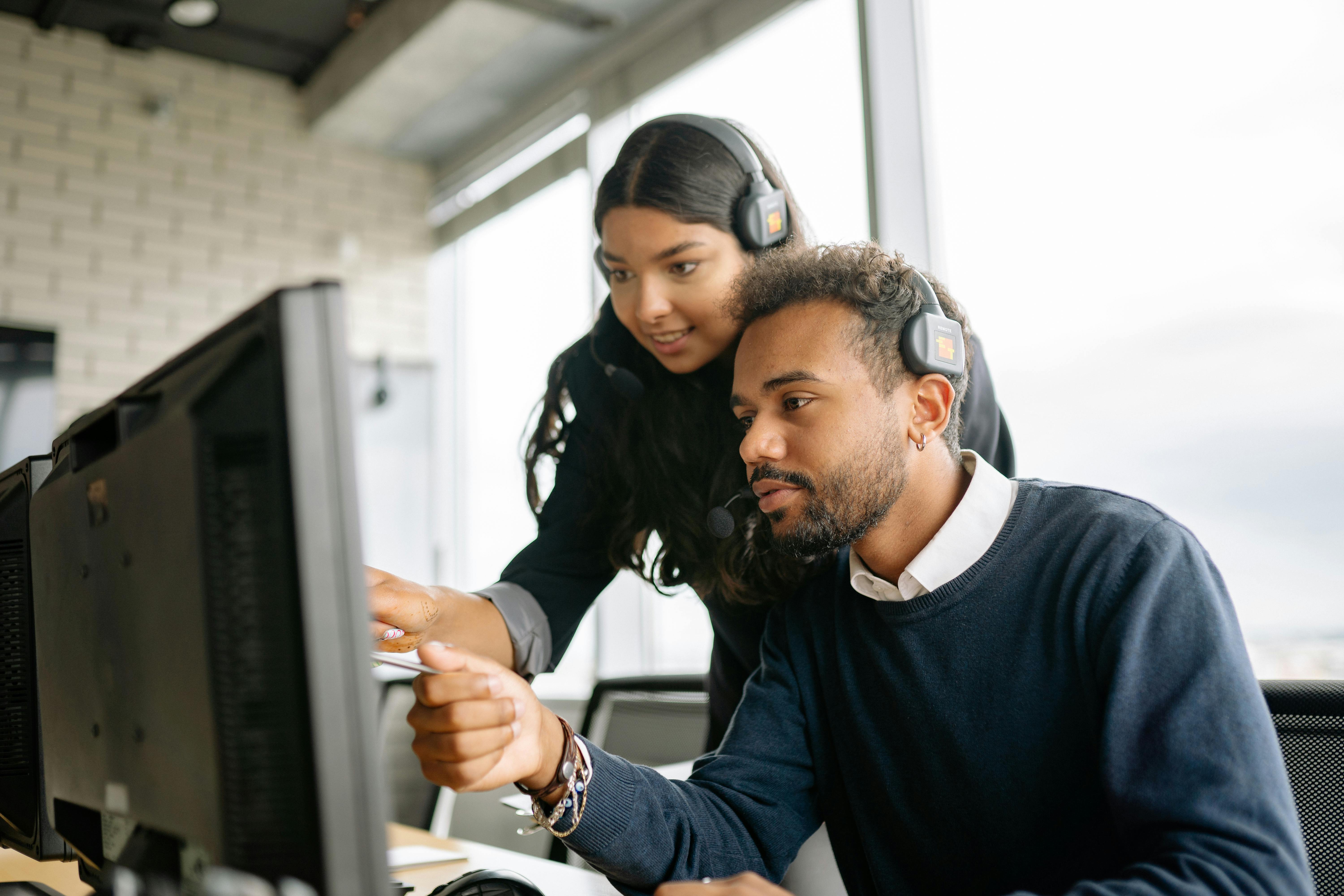 Two IT support specialists wearing headphones and discussing something on monitor